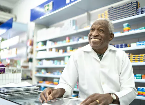 A smiling pharmacist behind the counter of a well-stocked pharmacy